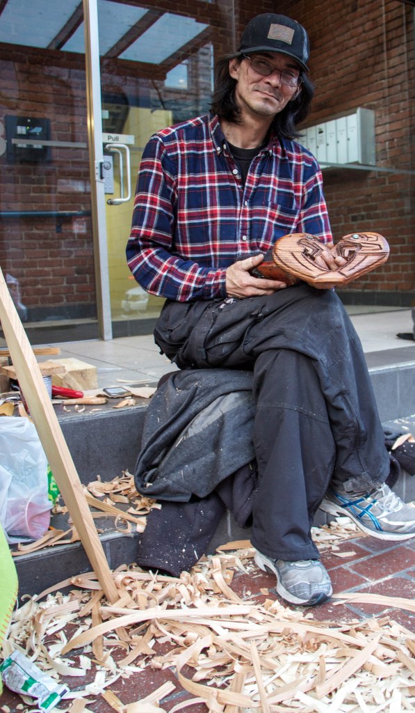 Aboriginal artist Darcy Coon carves a paddle in Gastown, where he sells his art to passersby. He holds a carving of two eagles forming a heart. Photo: Anna Dimoff